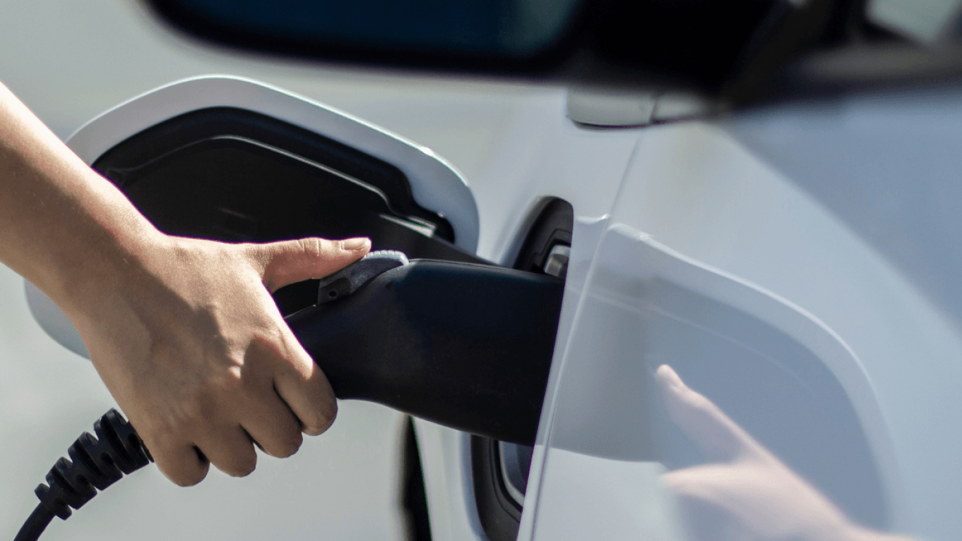 Nahaufnahme eines E-Autos beim Ladevorgang an einer Ladesäule – eine Hand steckt das Ladekabel in den Ladeanschluss des Fahrzeugs. Symbolbild für moderne E-Mobilität und Ladeinfrastruktur an der Tankstelle.