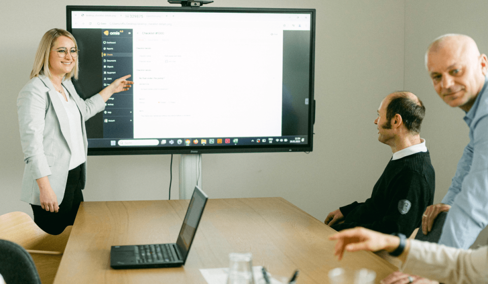 Mitarbeiterinnen und Mitarbeiter von omis bei einer Software-Präsentation im Meetingraum – eine Kollegin erklärt die Benutzeroberfläche des omis Tools auf einem großen Bildschirm.