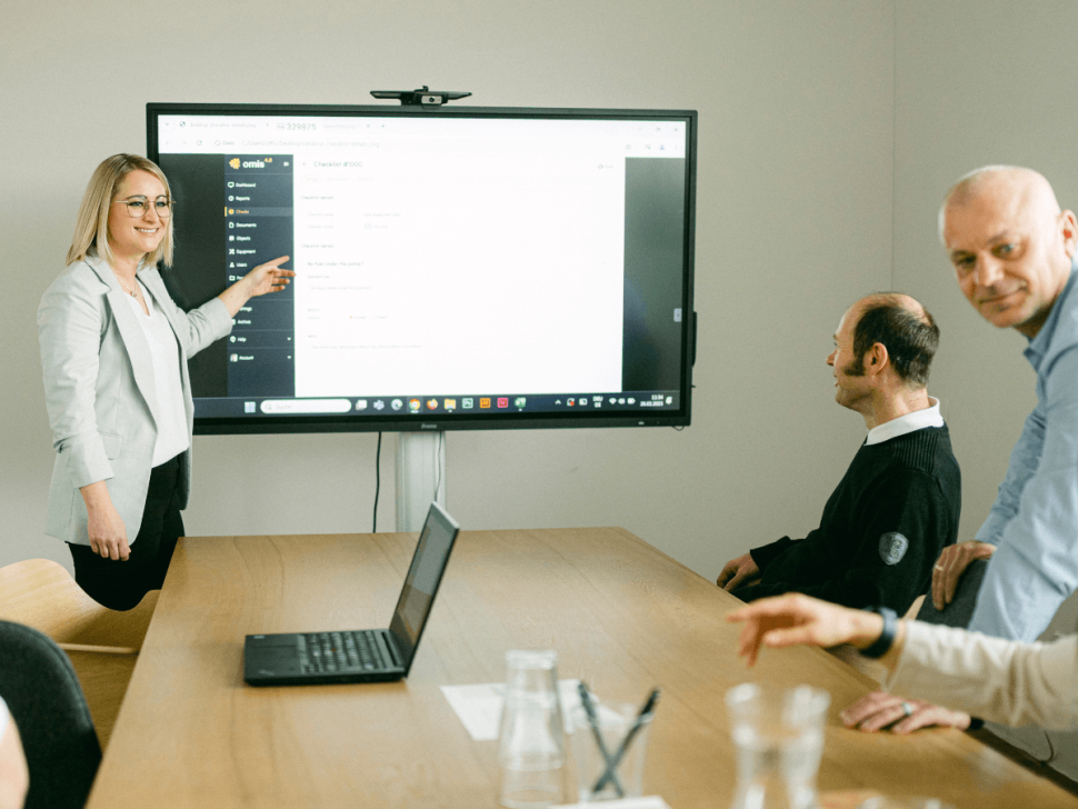 Mitarbeiterinnen und Mitarbeiter von omis bei einer Software-Präsentation im Meetingraum – eine Kollegin erklärt die Benutzeroberfläche des omis Tools auf einem großen Bildschirm.
