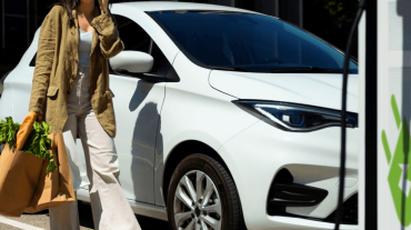 Frau mit Einkaufstüten und Smartphone vor einem weißen E-Auto an einer Ladesäule – Symbolbild für komfortables Laden während des Einkaufs an der Tankstelle.