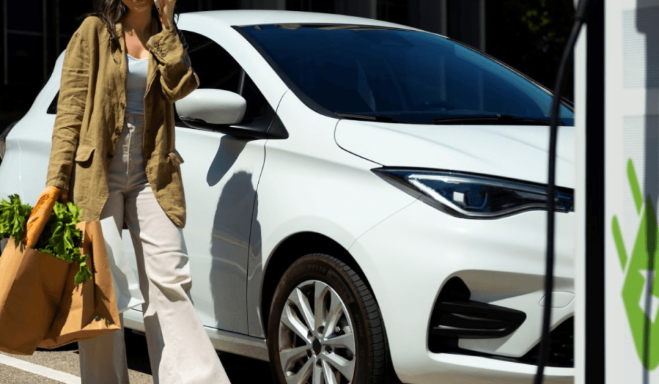 Frau mit Einkaufstüten und Smartphone vor einem weißen E-Auto an einer Ladesäule – Symbolbild für komfortables Laden während des Einkaufs an der Tankstelle.
