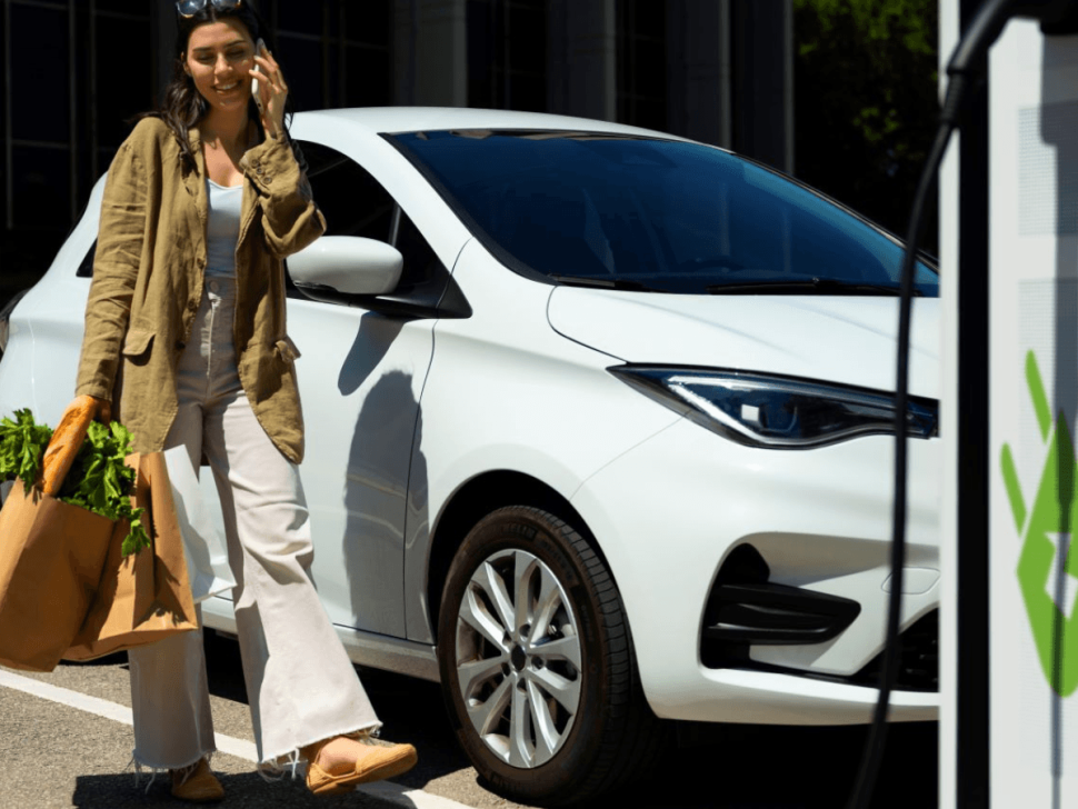 Frau mit Einkaufstüten und Smartphone vor einem weißen E-Auto an einer Ladesäule – Symbolbild für komfortables Laden während des Einkaufs an der Tankstelle.