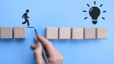 Symbolische Darstellung einer Lösung: Eine Hand zeichnet eine Brücke zwischen zwei Holzblöcken, um einer Figur den Weg zur Glühbirne – dem Symbol für eine Idee oder Innovation – zu ermöglichen.