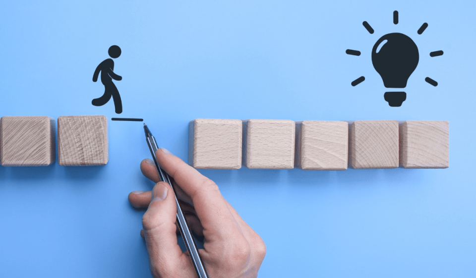 Symbolische Darstellung einer Lösung: Eine Hand zeichnet eine Brücke zwischen zwei Holzblöcken, um einer Figur den Weg zur Glühbirne – dem Symbol für eine Idee oder Innovation – zu ermöglichen.