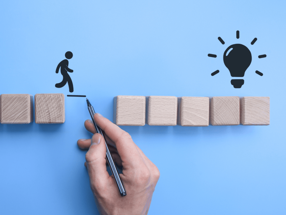 Symbolische Darstellung einer Lösung: Eine Hand zeichnet eine Brücke zwischen zwei Holzblöcken, um einer Figur den Weg zur Glühbirne – dem Symbol für eine Idee oder Innovation – zu ermöglichen.